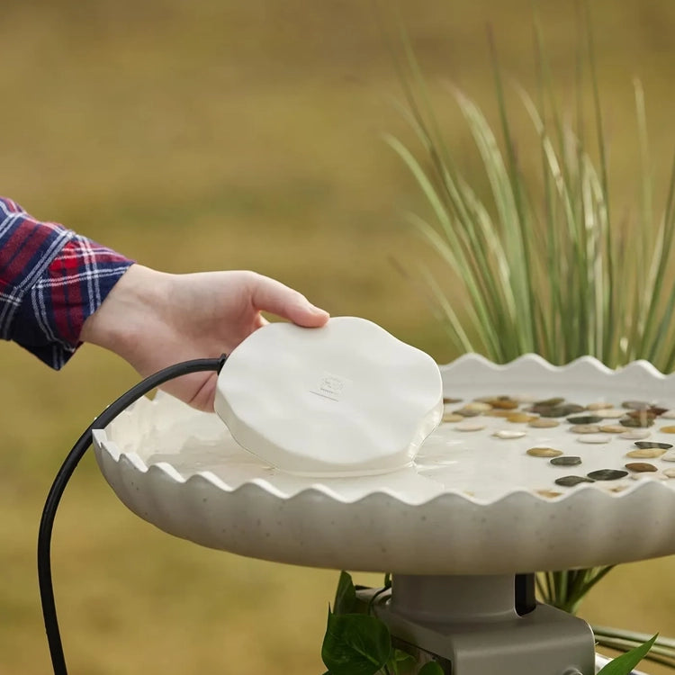 A hand lowers the Heated Rock Bird Bath De-icer into a scalloped bird bath, featuring decorative stones, to prevent water from freezing.