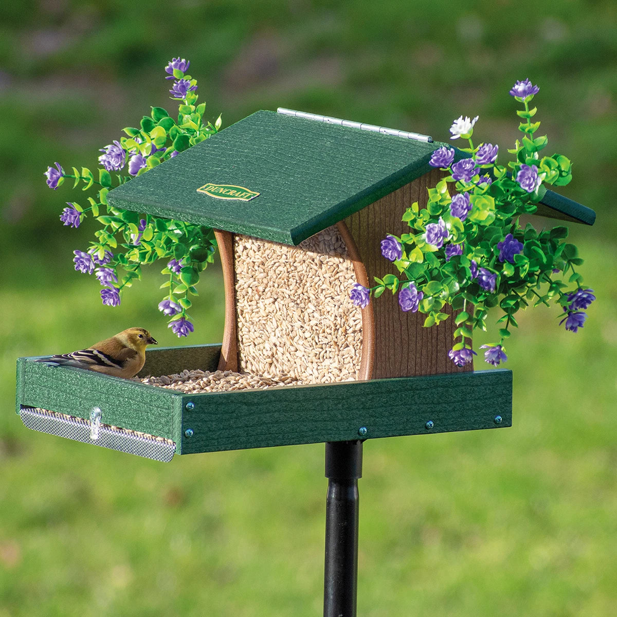 Duncraft Back to the Woods Cardinal Landing Tray with Pole features a green mounted feeder, goldfinch eating, and decorative flowers flanking the roof.