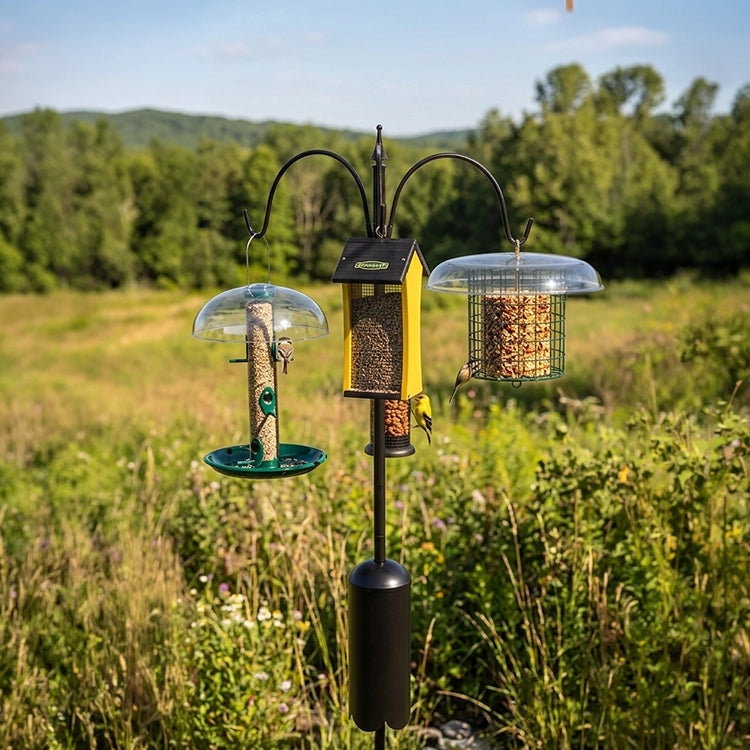 Three bird feeders on a stand in a natural setting with trees and grass.