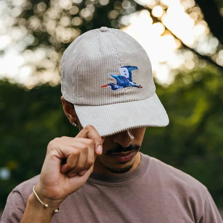 Young man outdoors, tipping the brim of an ivory Great Blue Heron Corduroy Hat, featuring an embroidered bird design, embodying an adventure-ready style.
