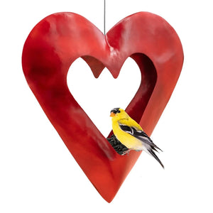 Fly Thru Red Heart Copper Bird Feeder with a heart-cutout, red finish; an American goldfinch feeds on sunflower seeds, showcasing the feeder's inviting design.