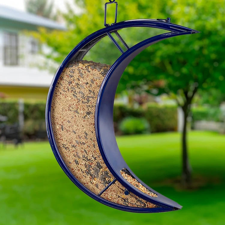 Over the Moon Fly-Thru Bird Feeder, crescent-shaped, glossy blue, filled with seeds, hanging outdoors, offers birds a spacious perch for feeding.