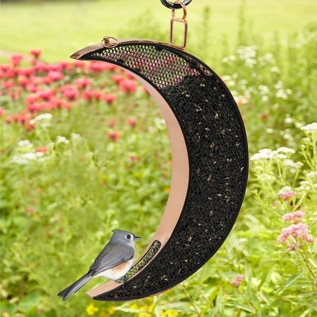 Over The Moon Copper Bird Feeder with mesh panels, showcasing its crescent shape, hangs in a garden with a songbird perched, enjoying seeds.