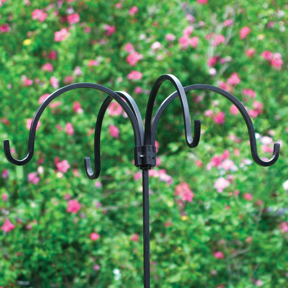 Adjustable Quad Bird Feeder Hanger made of black wrought iron, shown with flowers in the background, featuring four hangers and an adjustable height pole.