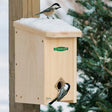 Duncraft Convertible Roost House with snow-dusted roof; two chickadees explore. Provides warm shelter in winter, converts to nesting box in spring.