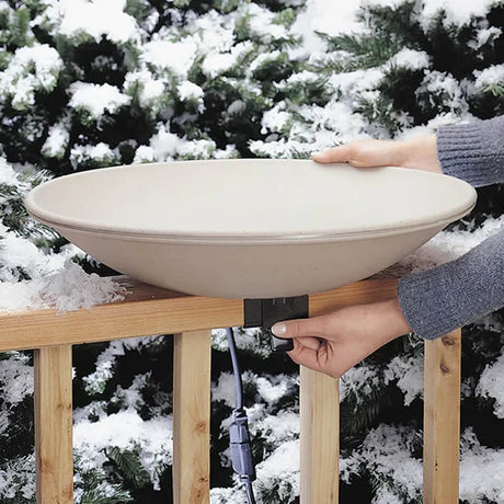 Hands adjust All-Seasons Heated Deck Mount Bath on a railing, showcasing a shallow dish design with a coaxial cable in a snowy winter setting.