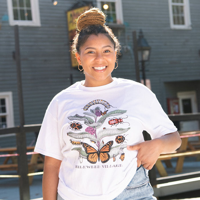 Woman smiling and pointing at her Milkweed Village T-Shirt, featuring a nature-inspired design by Diana Dee Tyler, printed on a 100% cotton, white Gildan heavyweight shirt.