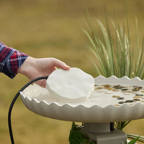 A hand lowers the Heated Rock Bird Bath De-icer into a scalloped bird bath, featuring decorative stones, to prevent water from freezing.