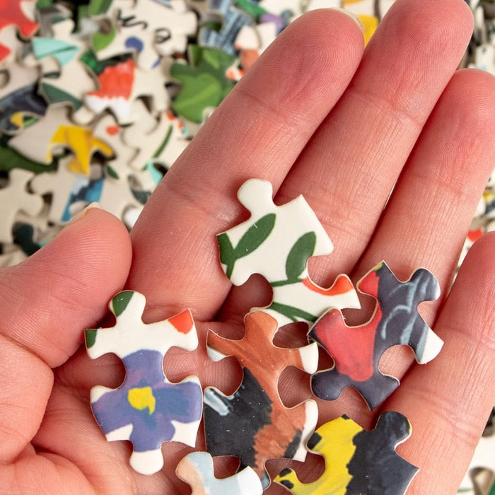 Hand holding a piece of the Feathered Friends 1,000 Piece Bird Jigsaw Puzzle, featuring hand-painted birds and species names, perfect for bird watchers and nature enthusiasts.