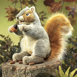 Gray Squirrel Hand Puppet sitting on a stump, holding an acorn with a playful expression and soft, bushy tail.