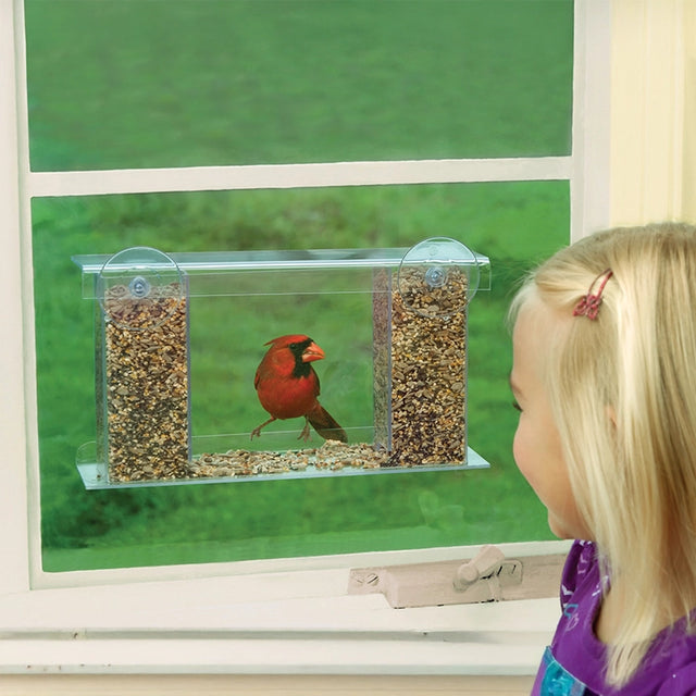 A clear window-mounted bird feeder filled with seeds, with a red cardinal perched on the front, while a young girl in a purple shirt looks out the window.