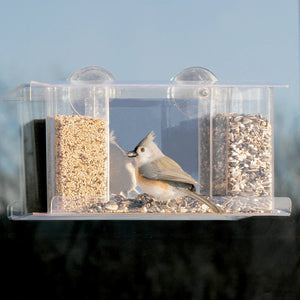 Duncraft Songbird Mirrored Window Feeder with a bird perched inside, eating seeds. The clear plastic feeder attaches to the window with two suction cups.