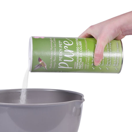 Hand pouring Perky-Pet® Pure Clear Hummingbird Nectar Powder, 24 oz., into a bowl, demonstrating its easy pour and mix feature for making nectar.