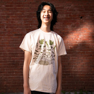 Man wearing Frog Metamorphosis T-Shirt featuring a graphic design of frog metamorphosis, standing in front of a brick wall.