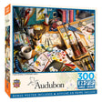 Audubon Creative Birding 300 pc. EZ-Grip Puzzle features art supplies and bird-themed items on a cluttered desk, ideal for bird lovers and families.