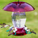 Jubilee Hummingbird Feeder with red weather guard, clear reservoir, and four white flower-shaped feeding ports, attracting birds. Includes S-hook and ant moat for easy hanging.