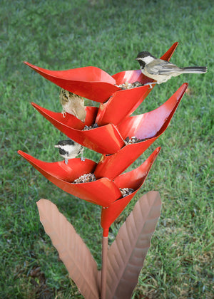 Heliconia Bird Feeder: A bird perched on a metallic red flower-shaped feeder designed to cascade seeds through each petal, enhancing backyard bird-watching.