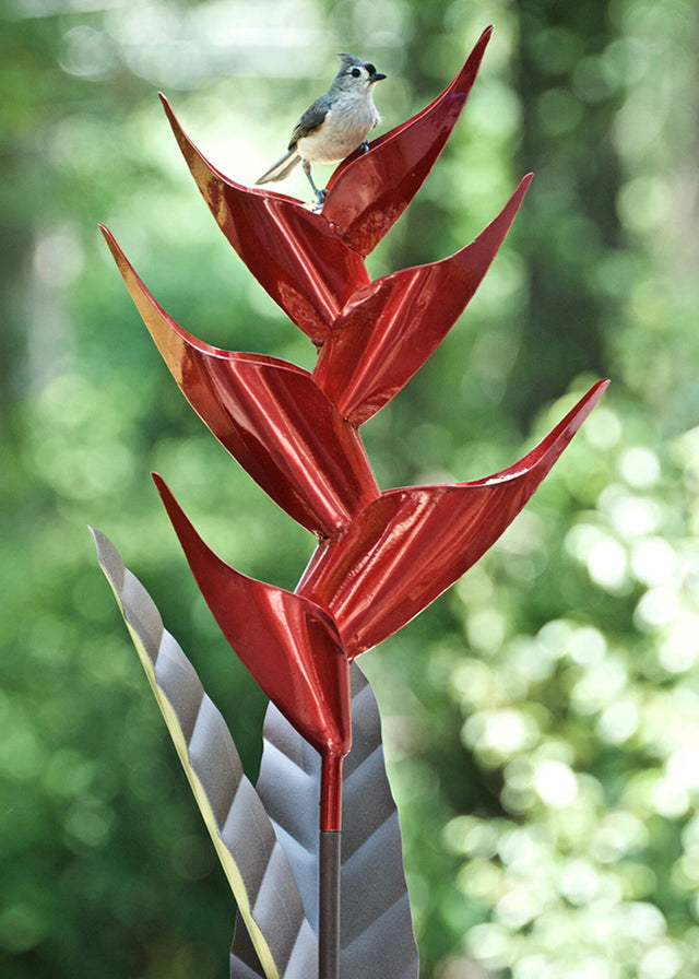 Heliconia Bird Feeder: A bird perches on a red, metallic plant-shaped feeder designed for cascading bird seed, featuring durable, all-metal construction.