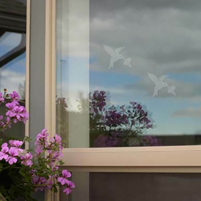 Hummingbird WindowAlert Decals displayed on a window with purple flowers nearby, designed to prevent bird collisions by reflecting ultraviolet light. Set of 4 decals.