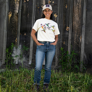 Hummingbirds T-Shirt: Woman in white shirt with colorful hummingbirds, blue jeans, and hat standing outdoors near a fence.