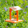 Duncraft Sheltered Oriole Jelly Feeder with orange recycled plastic and clear roof, featuring an oriole perched, feeding from the metal dish. Includes hanging hook and chain.