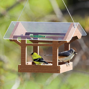 Duncraft Covered Bridge Platform Feeder with a yellow bird perched, featuring a clear protective roof and fly-through openings for feeding on all sides.