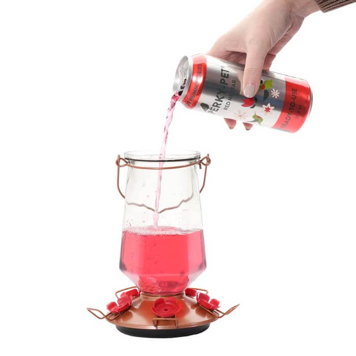 Hand pouring Perky-Pet® Ready-To-Use Red Hummingbird Nectar, 16 oz., from an aluminum can into a glass container.