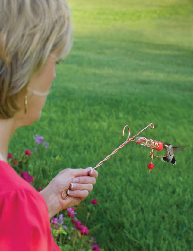 Whimsy Wand Hummer Feeder: a woman holding a glass tube hummingbird feeder attached to a copper wand, featuring a red bead accent.