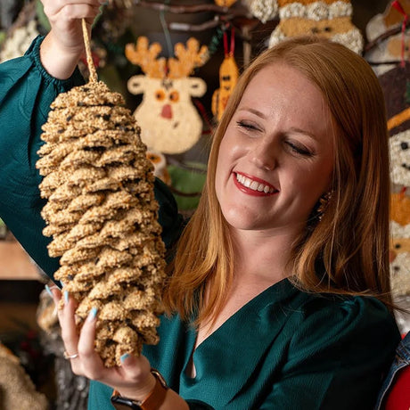 Woman holding Giant Pine Cone for The Birds, a seed-coated treat for birds, amidst festive decorations, showcasing its size and holiday gift appeal.