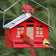 Squirrel Be Gone Country bird feeder: red metal house design with cardinal weathervane, featuring Northern Cardinal and Goldfinch on weight-activated perch against green foliage.