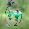 Balance 2 Hummingbird Feeder, Green, with two red flower feeding tubes and a handblown recycled glass reservoir, hanging on a wire with a hummingbird perched.