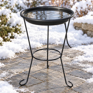 Black heated bird bath with clear water on a three-legged metal stand, surrounded by snow on a stone patio