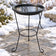 Black heated bird bath with clear water on a three-legged metal stand, surrounded by snow on a stone patio