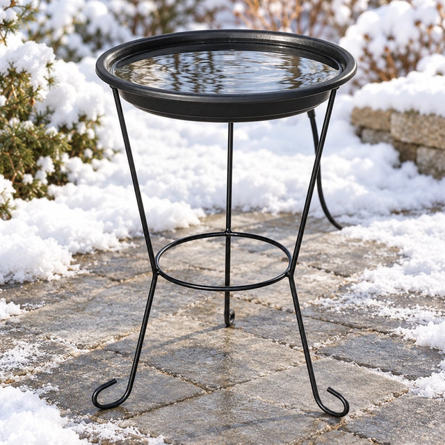 Black heated bird bath with clear water on a three-legged metal stand, surrounded by snow on a stone patio