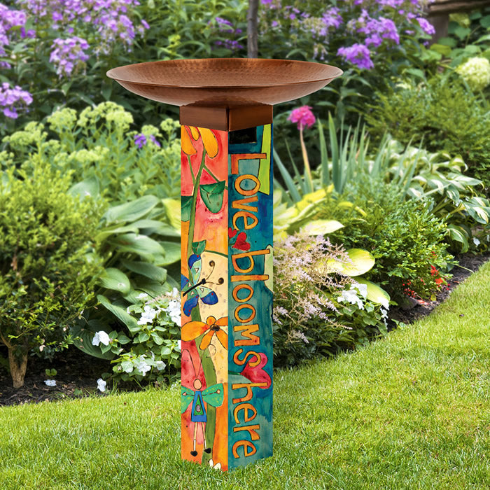 Love Garden Bird Bath with copper-plated topper, vibrant floral illustrations, and “Love blooms here” text, standing in a garden, 18-inch diameter basin on a sturdy PVC post.