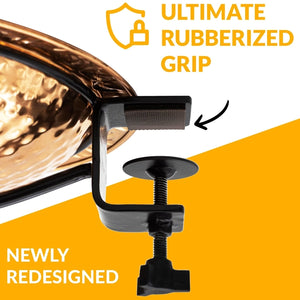 A close-up promotional shot of a black clamp-style cup holder with a rubberized grip, shown attached to the rim of a hammered copper pot. Text overlays read 'ULTIMATE RUBBERIZED GRIP' and 'NEWLY REDESIGNED' on a white and orange background.