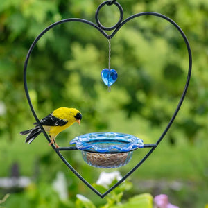 A heart-shaped black metal bird feeder with a blue glass seed tray and a hanging blue glass heart ornament, shown outdoors with a yellow bird perched on the left against a blurred green garden background.