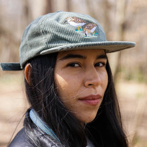 Bird Collective Least Sandpipers Chunky Corduroy Hat worn by a smiling woman, showcasing the bird design and adjustable strap for outdoor adventures.