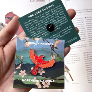 Hand holding the Bird Collective Northern Cardinal or Eastern Bluebird enamel pin attached to a green card.