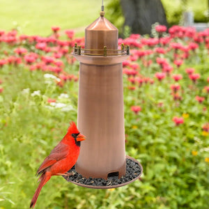 Copper Lighthouse Bird Feeder with a red Northern Cardinal perched on the seed tray, surrounded by soft-focus garden foliage and pink flowers.