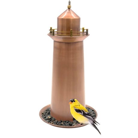 Copper Lighthouse Bird Feeder with conical top and railing, filled with sunflower seeds. An American goldfinch perches on the tray, feeding.