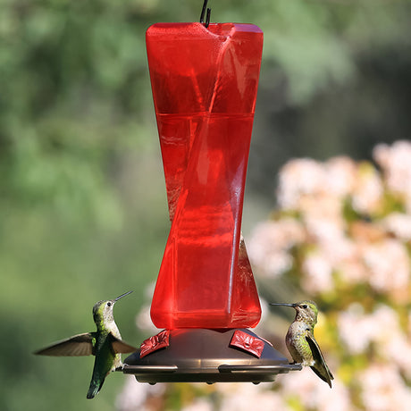 Mirage Hummingbird Feeder with two hummingbirds feeding at red flower ports on an antique copper finish base, featuring a see-through plastic reservoir.