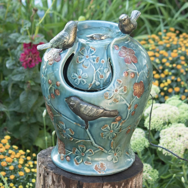 Blue Verdigris Songbird Fountain with two birds perched on the rim, surrounded by antiqued flowers and branches, illuminated by LED lights, creating a tranquil atmosphere.