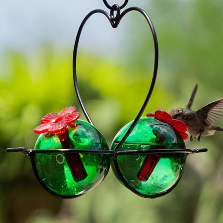 Hanging Droplet Heart 2 Hummingbird Feeder with green glass nectar sphere and heart-shaped black wire holder, featuring a hummingbird in flight nearby.