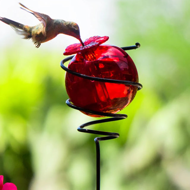 Droplet Spiral on a Stake Hummingbird Feeder, Red, features a glass nectar sphere with a flower-shaped port, held by a metal stake, attracting hummingbirds.