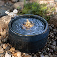 Elloise LED Bird Fountain featuring a distressed white bird statue on a navy blue, embossed plaid ceramic fountain with LED lights and natural pebbles.