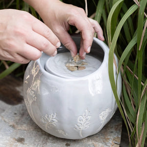 Hands placing pebbles into the Wildflower Fountain, a gray ceramic orb with botanical patterns, designed for a tranquil tabletop water feature.