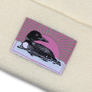 Bird Collective Loon Cuffed Beanie, Ivory, featuring a woven patch of a loon on a white knitted surface.