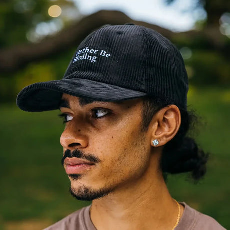 Young man wearing the I'd Rather Be Birding black corduroy hat with adjustable strap, showcasing its relaxed, unstructured fit in a park setting.
