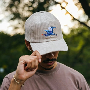 Young man outdoors, tipping the brim of an ivory Great Blue Heron Corduroy Hat, featuring an embroidered bird design, embodying an adventure-ready style.